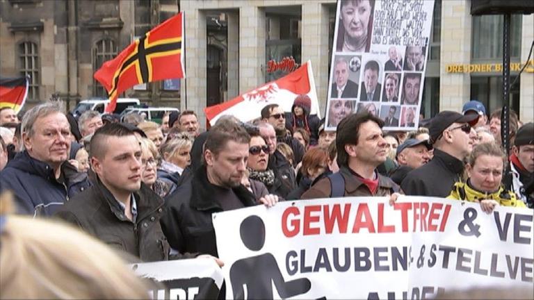 "Sachsen zwischen Mauerfall und Rechtspopulismus - Eine Spurensuche": Eine Gruppe von Menschen demonstriert auf der Straße. Einige halten Banner und Fahnen in der Hand.