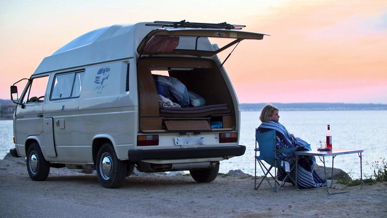 "Die neue Lust am Campen - Vier Räder statt vier Sterne": Neben einem Campingbus sitzt Eva Mühlenbäumer am Meeresufer an einem Campingtisch, auf dem eine Flasche Wein und zwei Gläser stehen.