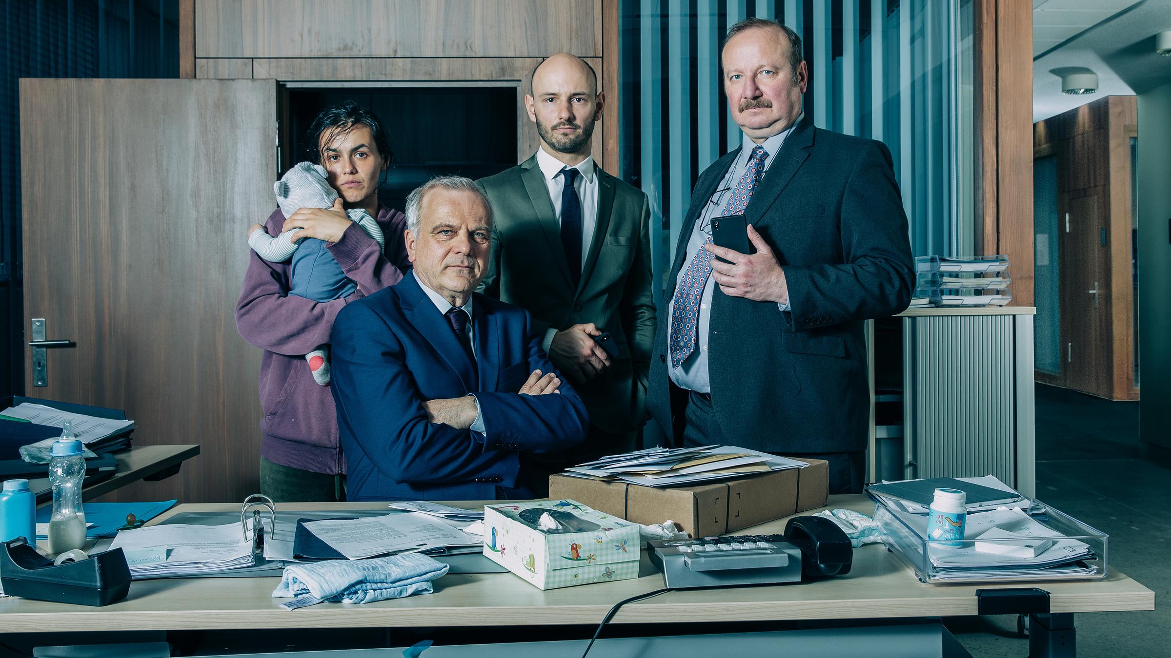 "Eichwald, MdB: Männer können alles":  Eichwald (Bernard Schütz) sitzt hinter einem mit Ordner, Taschentuchbox, Paket, Teafilmabroller und Ablagekorb belegtem Schreibtisch. Direkt hinter ihm steht Sebastian Grube (Leon Ulrich) mit verschränkten Händen. Neben ihm steht Berndt Engemann (Rainer Reiners) mit Handy in der linken Hand. Vor der geöffneten Bürotür steht Julia Schleicher (Lucie Heinze) und hält ein Baby in den Armen. Ihre Hand stützt den Kopf des Babys.