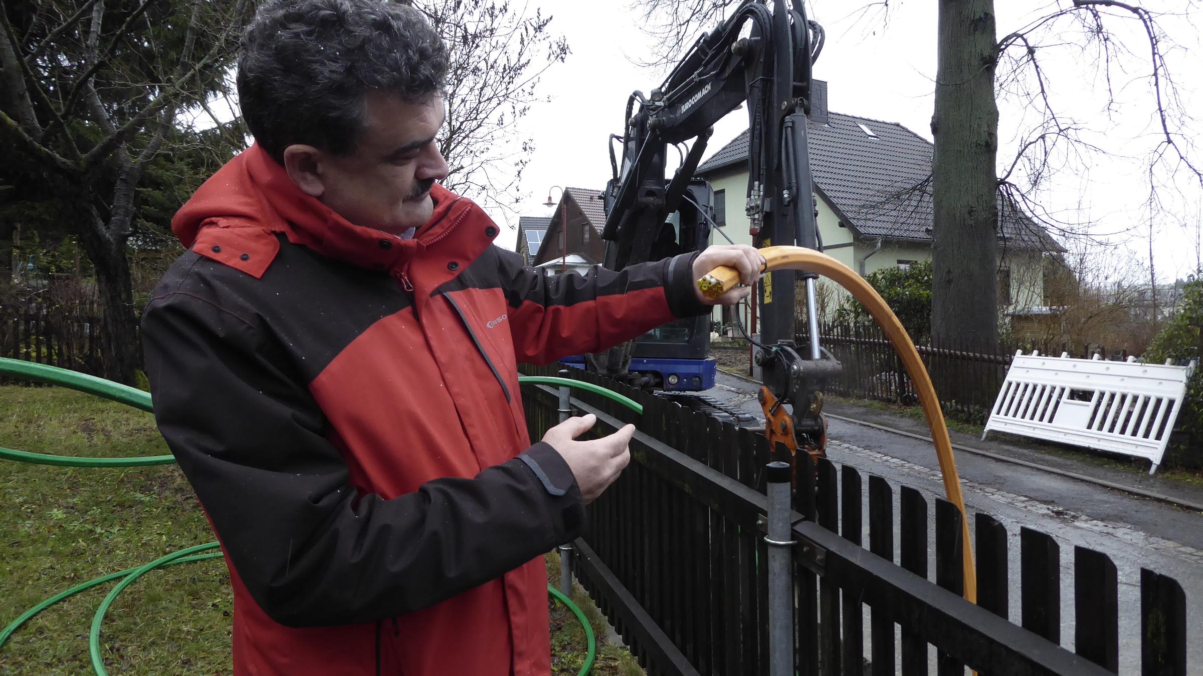 "WISO-Dokumentation: Kunden in der Warteschleife - Die langen Leitungen von Telekom & Co.": Bürgermeister Weigt steht mit einem Telekommunikationskabel in der Hand an einem Holzzaun. Im Hintergrund sind ein Bagger und Wohnhäuser zu sehen.