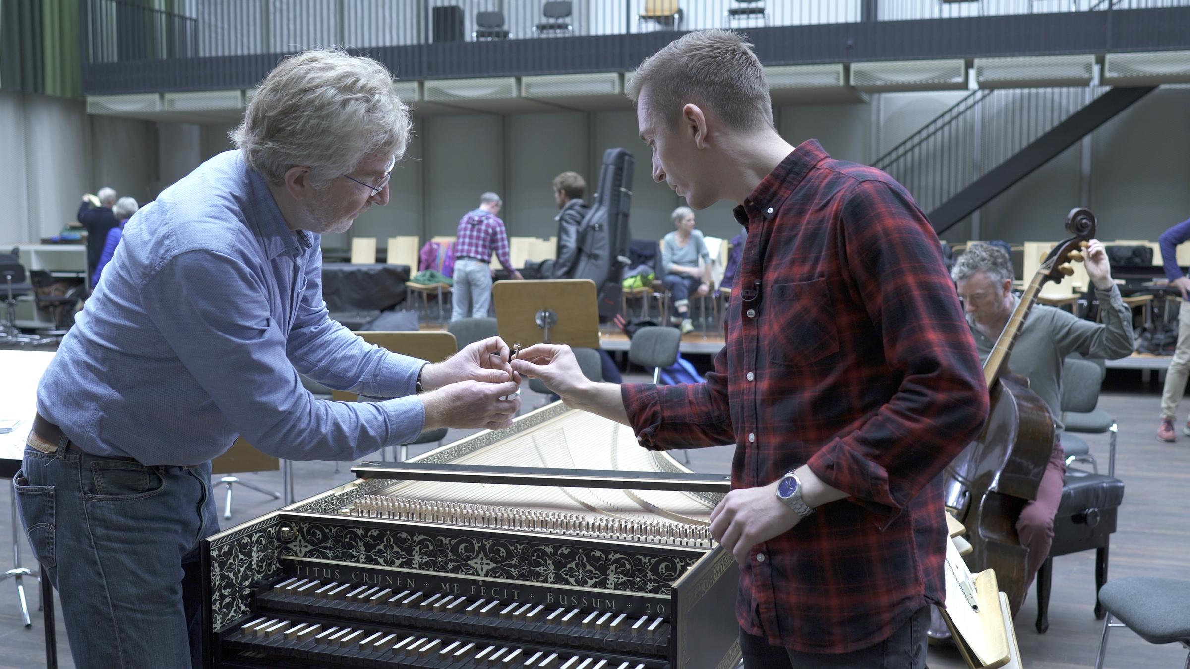 "Bühnenzauber - Hinter den Kulissen der Berliner Staatsoper": Eric Mayer (r.) und ein Mitarbeiter der Berliner SStaatsoper stehen über ein Piano gebeugt in einem Probenraum. Im Hintergrund läuft ein Mannn mit einem Geigenkasten auf dem Rücken vorbei. 