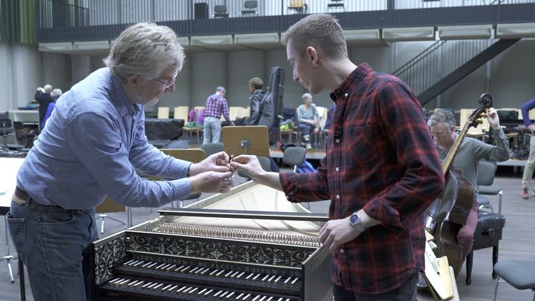 "Bühnenzauber - Hinter den Kulissen der Berliner Staatsoper": Eric Mayer (r.) und ein Mitarbeiter der Berliner SStaatsoper stehen über ein Piano gebeugt in einem Probenraum. Im Hintergrund läuft ein Mannn mit einem Geigenkasten auf dem Rücken vorbei. 