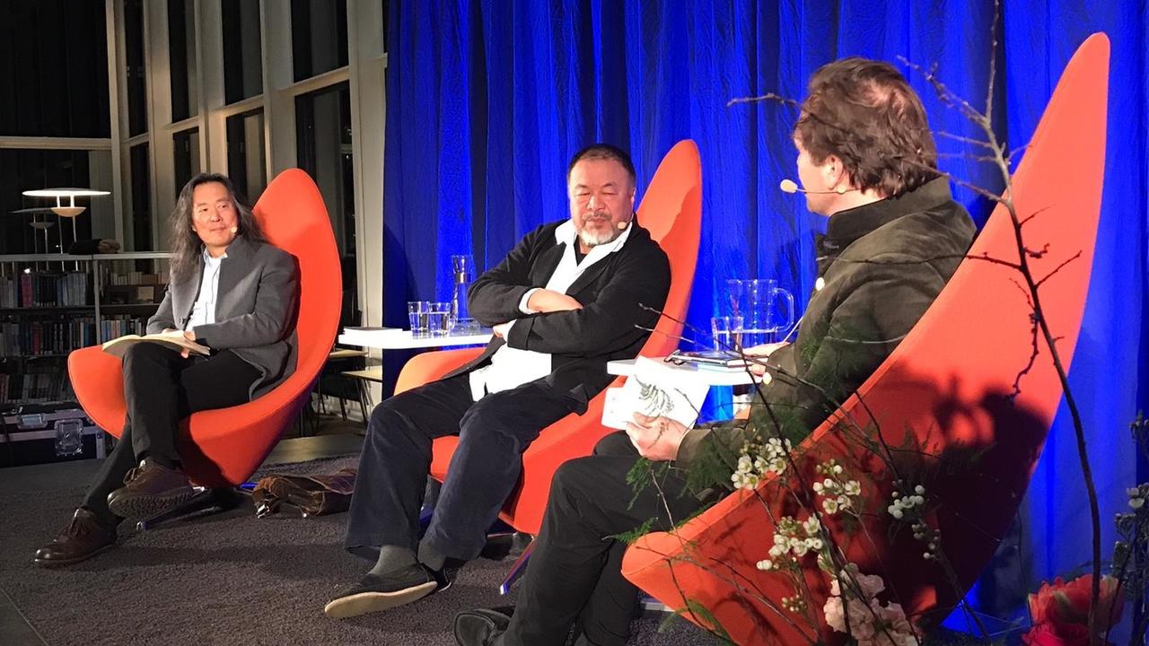 "Alles ist Kunst, alles ist Politik – Ai Weiwei in Düsseldorf":  Auf dem Bild sind drei Männer auf einem Podium zu sehen. Alle drei sitzen auf roten Stühlen mit spitzer, hoher Lehne. Der Moderator ist nur in Rückenansicht zu sehen. In der Mitte sitzt der Künstler Ai Weiwei und verschränkt die Arme. Außen links im Bild sitzt der chinesische Schriftsteller Yang Lian.