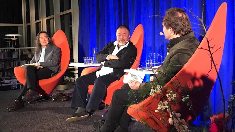 "Alles ist Kunst, alles ist Politik – Ai Weiwei in Düsseldorf":  Auf dem Bild sind drei Männer auf einem Podium zu sehen. Alle drei sitzen auf roten Stühlen mit spitzer, hoher Lehne. Der Moderator ist nur in Rückenansicht zu sehen. In der Mitte sitzt der Künstler Ai Weiwei und verschränkt die Arme. Außen links im Bild sitzt der chinesische Schriftsteller Yang Lian.
