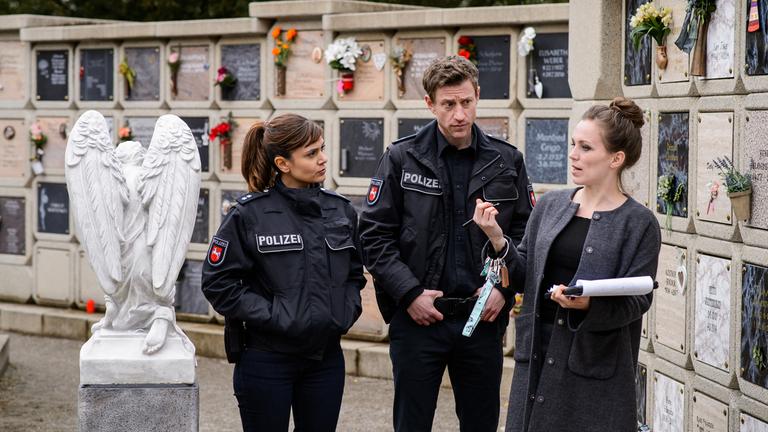 „Friesland – Asche zu Asche“: Süher Özlügül (Sophie Dal, l.) und Henk Cassens (Maxim Mehmet, M.) stehen vor einer Urnenwand und befragen Friedhofsverwalterin Mina Wimmer (Franziska Wulf, r.), die in einem  Arm einen Block und am anderen Arm einen Ring mit vielen Schlüsseln trägt.