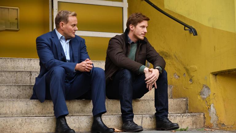 “Schwartz & Schwartz - Der Tod im Haus“: Andi (Devid Striesow) und Mads Schwartz (Golo Euler) sitzen beide auf einer Treppe. Die Brüder befinden sich vor einer Eingangstür und schauen nachdenklich in die Ferne.