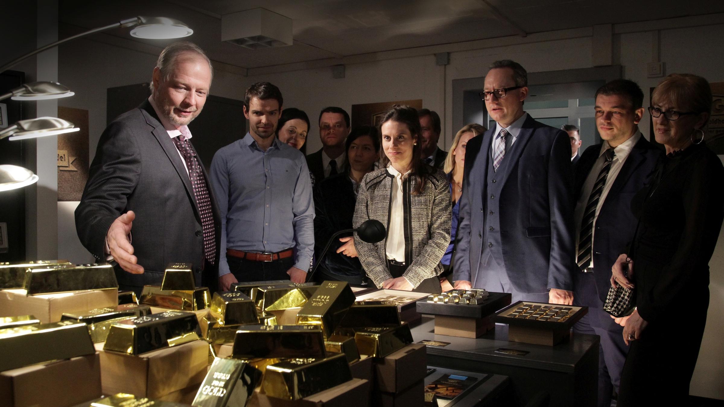 "Aktenzeichen XY-Spezial: Vorsicht, Betrug!": Auf einem Tisch liegen sehr viele Goldbarren. Ein Mann steht im Vordergrund und deutet mit der Hand auf die Barren. Dahinter sieht man mehrere Personen, die freudig auf das Gold schauen.