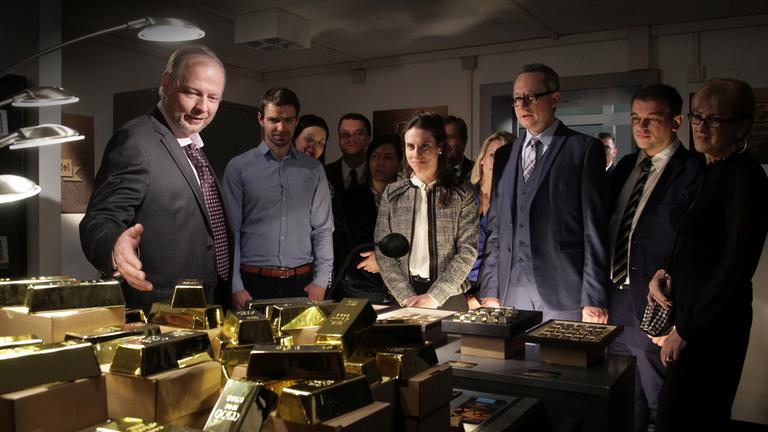 "Aktenzeichen XY-Spezial: Vorsicht, Betrug!": Auf einem Tisch liegen sehr viele Goldbarren. Ein Mann steht im Vordergrund und deutet mit der Hand auf die Barren. Dahinter sieht man mehrere Personen, die freudig auf das Gold schauen.