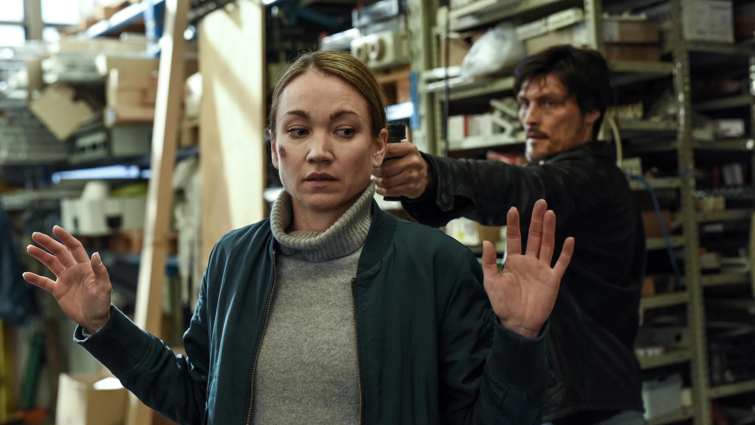„Sarah Kohr - Das verschwundene Mädchen“: Sarah (Lisa Maria Potthoff) steht mit erhobenen Händen in einem Lagerraum. Hinter ihr befindet sich Egor (Stipe Erceg), der ihr eine Waffe an den Kopf hält.