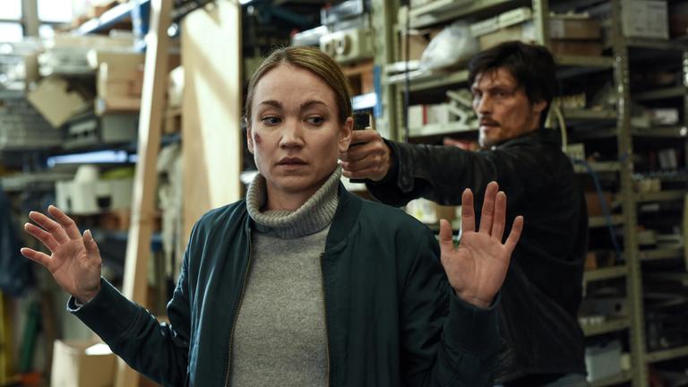 „Sarah Kohr - Das verschwundene Mädchen“: Sarah (Lisa Maria Potthoff) steht mit erhobenen Händen in einem Lagerraum. Hinter ihr befindet sich Egor (Stipe Erceg), der ihr eine Waffe an den Kopf hält.