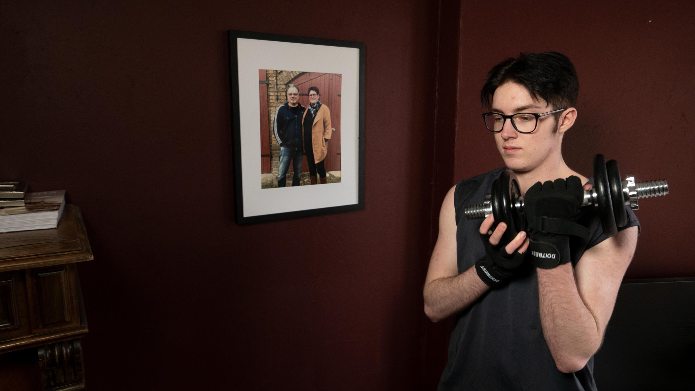 "37°: Glücklich erst als Junge - Wenn aus der Tochter ein Sohn wird": René steht mit einer Hantel in der Hand im Zimmer vor einem Foto seiner Eltern.