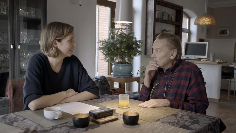 "Helmut Berger, meine Mutter und ich": Helmut Berger und Valesca Peters sitzen sich über Eck an einem Esstisch in einem großen Wohnraum gegenüber. Sie lächelt ihn an. Er blickt nachdenklich zu ihr.