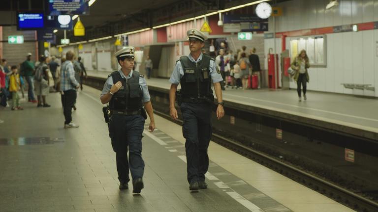 "Achtung Polizei! - Willkür, Pannen, Personalnot": Die Polizisten Kelly Schuster und Sebastian Wolz laufen an einem Bahngleis entlang. Im Hintergrund sind Menschen zu sehen.
