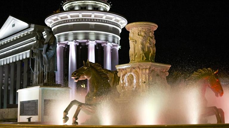 "Bigger Than Life": Ein Ensemble von vermeintlich historischen Monumenten wird nachts von Scheinwerfern erhellt: im Vordergrund ein Springbrunnen mit zwei Pferde-Skulpturen, links dahinter das Denkmal zweier antiker Männer, eine Säulen-Rotunde und eine Tempelfassade im griechischen Stil.