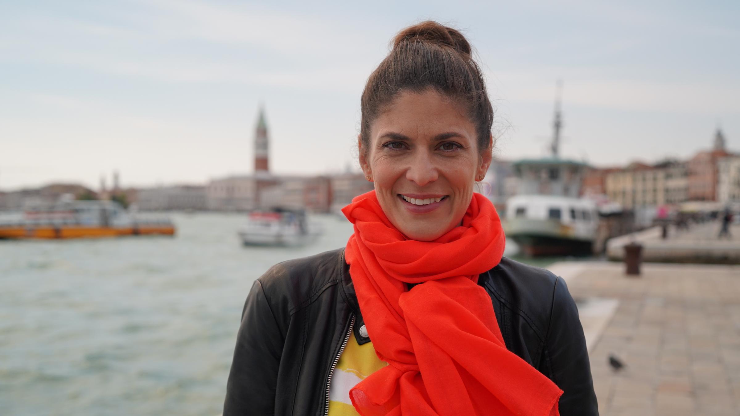 "aspekte - on tour: Biennale Venedig": Katty Salié  steht vor einer malerischen Kulisse in Venedig und lächelt in die Kamera.