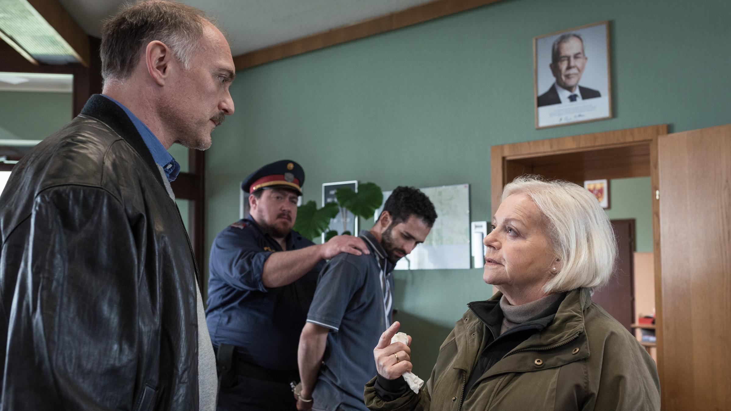 "Grenzland - Landkrimi": Achmet (Hassan Akkouch) wird von Hans Boandl (Christoph Krutzler) abgeführt. Vor ihnen im Zimmer stehen sich Elfriede Jandrasits (Brigitte Kren) und Dr. Schneider (Markus Schleinzer) gegenüber und unterhalten sich.