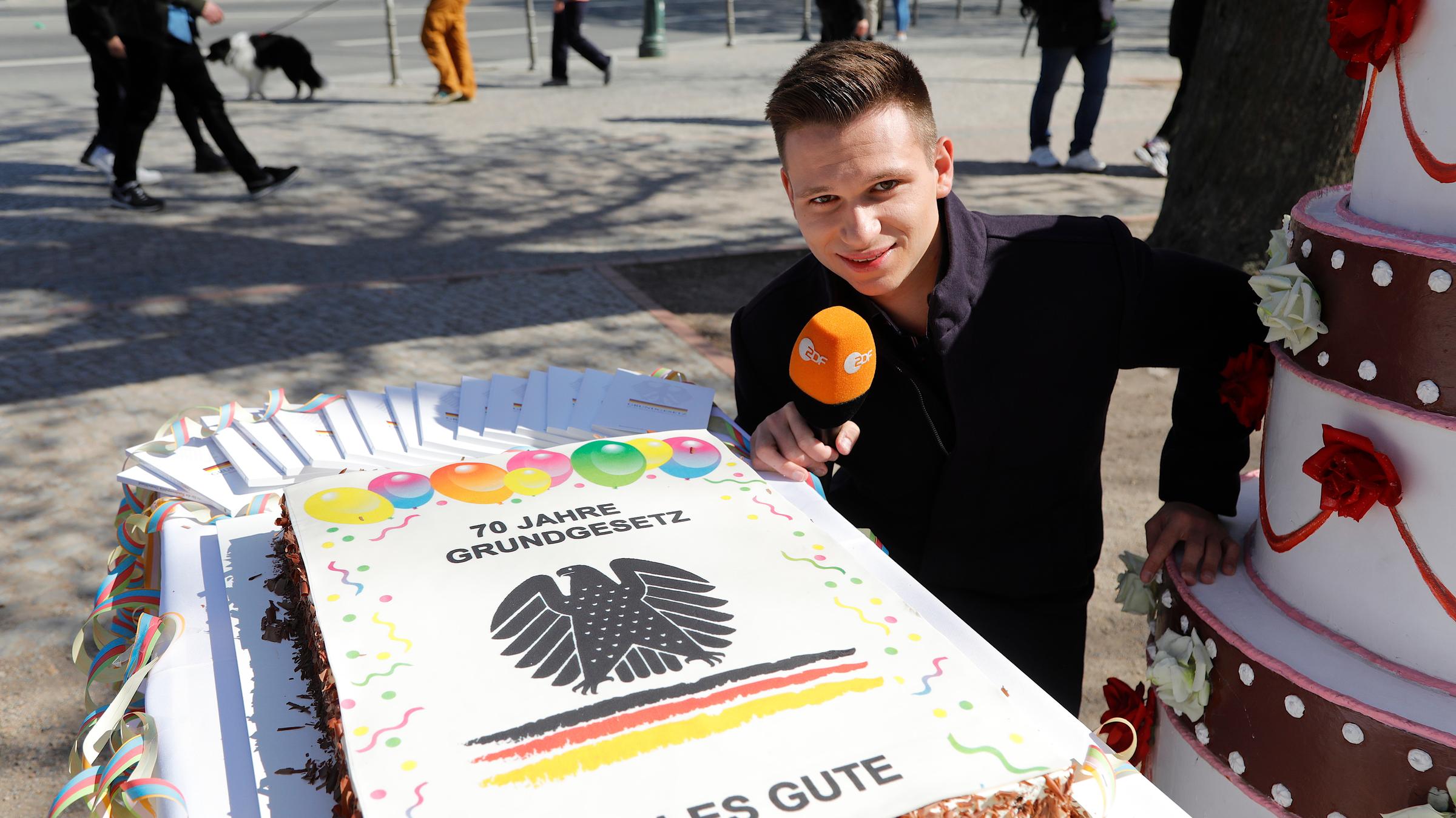 "ZDFzeit: 70 Jahre Grundgesetz - Die beste Verfassung der Welt?": Comedian Fabian Köster steht mit ZDF-Mikro auf einem belebten Platz an einer großen Geburtstagstorte mit Bundesadler und der Aufschrift "70 Jahre Grundgesetz. Alles Gute". Neben ihm steht eine ihn überragende mehrstöckige Deko-Torte, auf die er sich stützt.