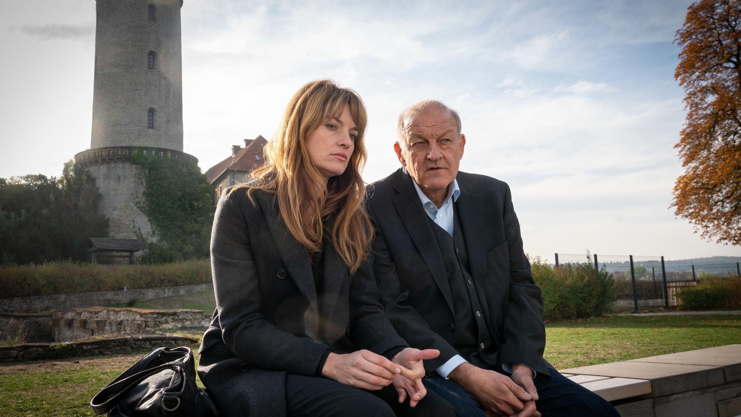 "Wilsberg - Ins Gesicht geschrieben":  Alex Holtkamp (Ina Paule Klink) sitzt mit traurigem Blick neben Georg Wilsberg (Leonard Lansink) auf einer Mauer. Im Hintergrund sieht man einen Turm der Sparrenburg.