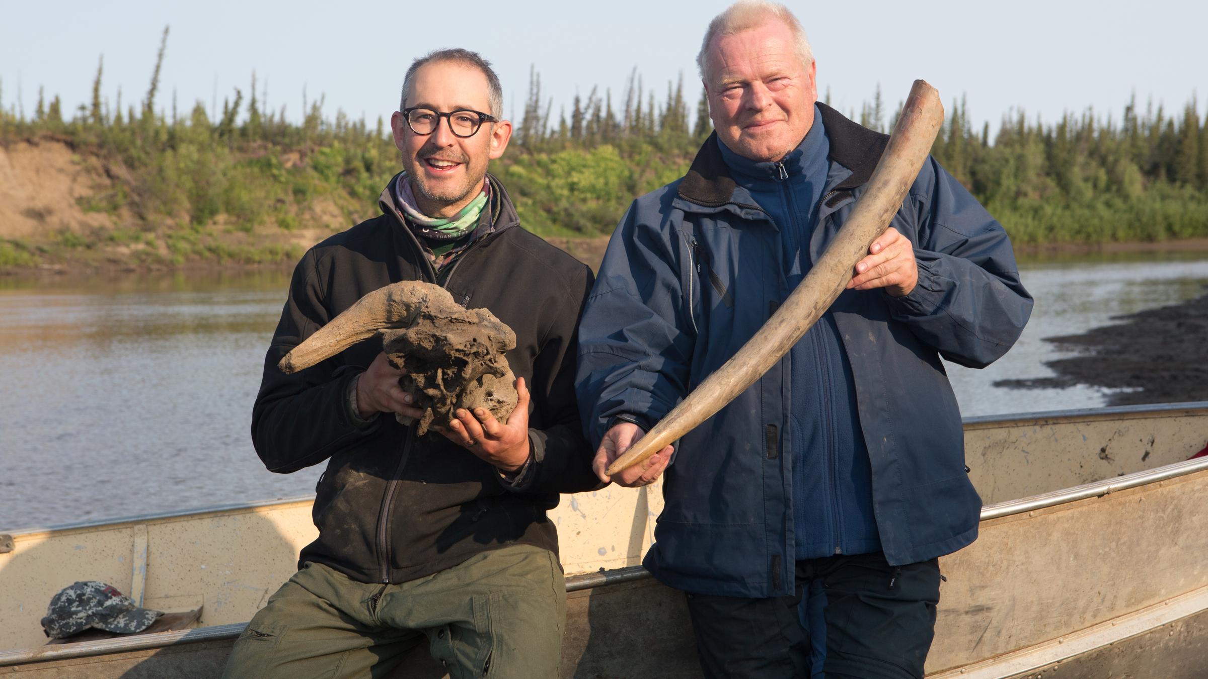 "Terra X: Das Eiszeiträtsel - Warum starben die Riesentiere aus?": Der Mammut-Experte Dick Mol und der Paläontologe Grant Zazula stehen nebeneinander an ein Ruderboot gelehnt an einem Fluss. Zazula hält einen Mammutstoßzahn in die Kamera, Mol den Schädel eines eiszeitlichen Moschusochsen.