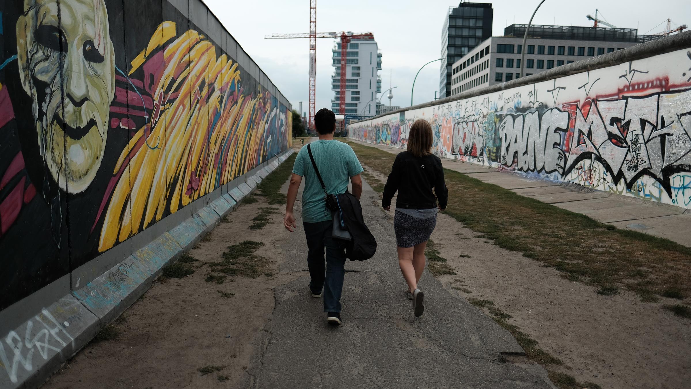 "ZDF.reportage: Post von drüben - Von grenzenloser Freundschaft": Ein Mann und eine Frau, beide mit dem Rücken zur Kamera, laufen  zwischen zwei Mauer-Elementen an der East-Side-Gallery.