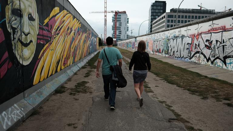 "ZDF.reportage: Post von drüben - Von grenzenloser Freundschaft": Ein Mann und eine Frau, beide mit dem Rücken zur Kamera, laufen  zwischen zwei Mauer-Elementen an der East-Side-Gallery.