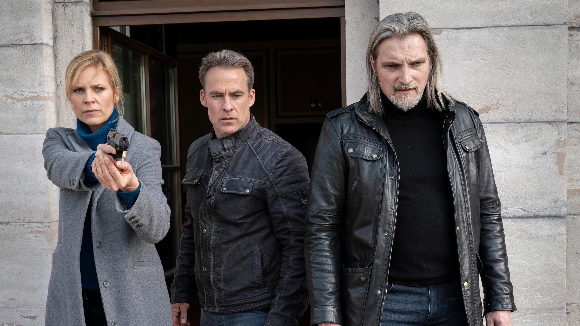 "Der vierte Mann": Ina Zimmermann (Melanie Marschke), Jan Maybach (Marco Girnth) und Carl Ribarski (Stefan Jürgens) stehen vor einem grauen Gebäude. Ina hat die Waffe gezückt. Alle drei schauen in die gleiche Richtung.
