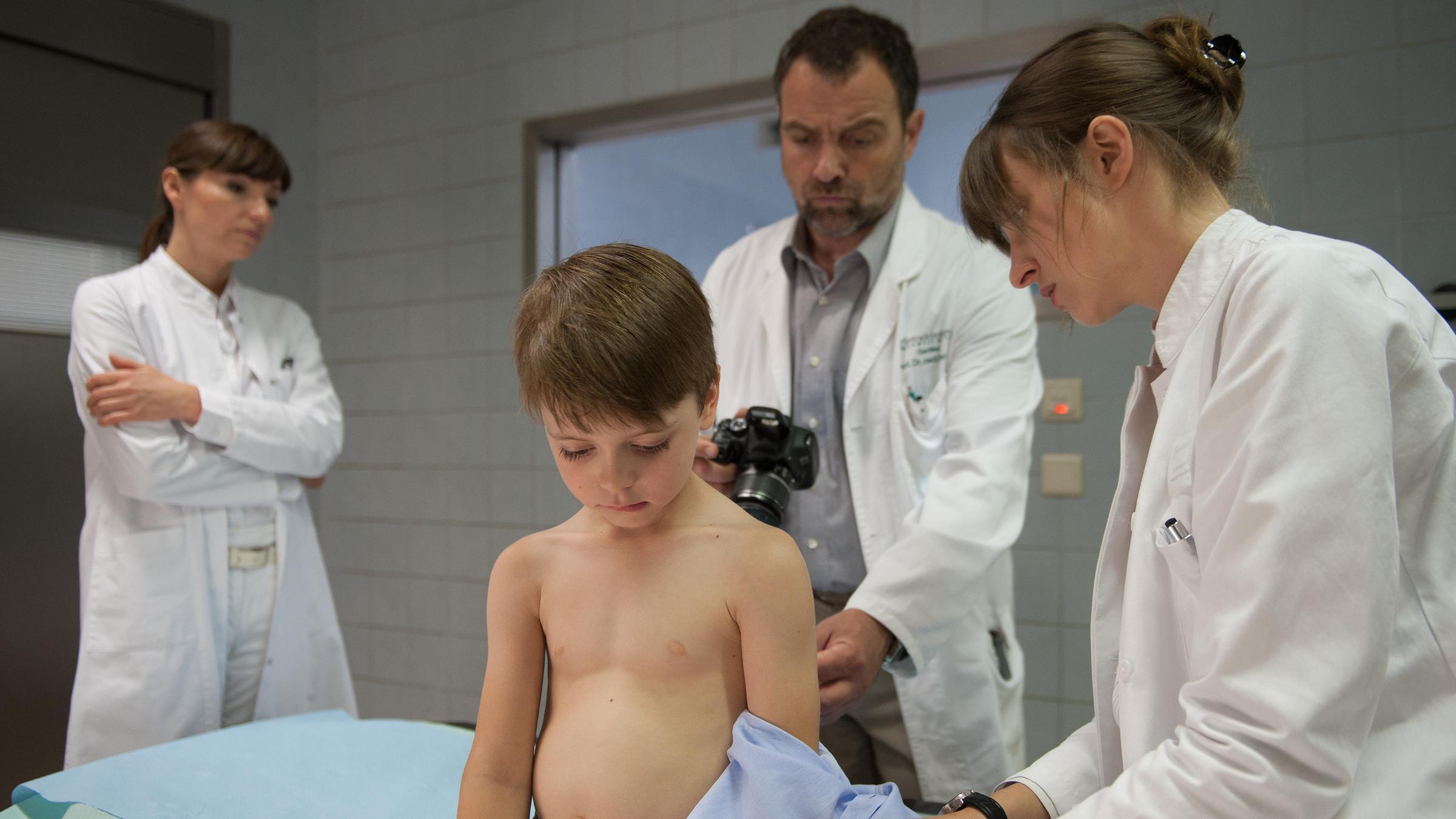 "Stumme Schreie": Dr. Bremer (Juergen Maurer) und Dr. Friedrich (Natalia Belitski) untersuchen den kleinen Paul (Elias), der vor ihnen auf der Behandlungsliege sitzt und unter sich schaut. Dr. Kern (Natascha Paulick) steht etwas abseits. Sie hat die Arme vor der Brust verschränkt und sieht ins Leere.
