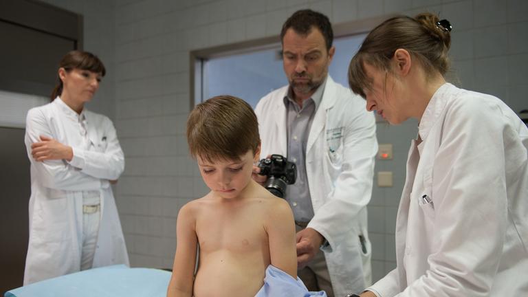 "Stumme Schreie": Dr. Bremer (Juergen Maurer) und Dr. Friedrich (Natalia Belitski) untersuchen den kleinen Paul (Elias), der vor ihnen auf der Behandlungsliege sitzt und unter sich schaut. Dr. Kern (Natascha Paulick) steht etwas abseits. Sie hat die Arme vor der Brust verschränkt und sieht ins Leere.