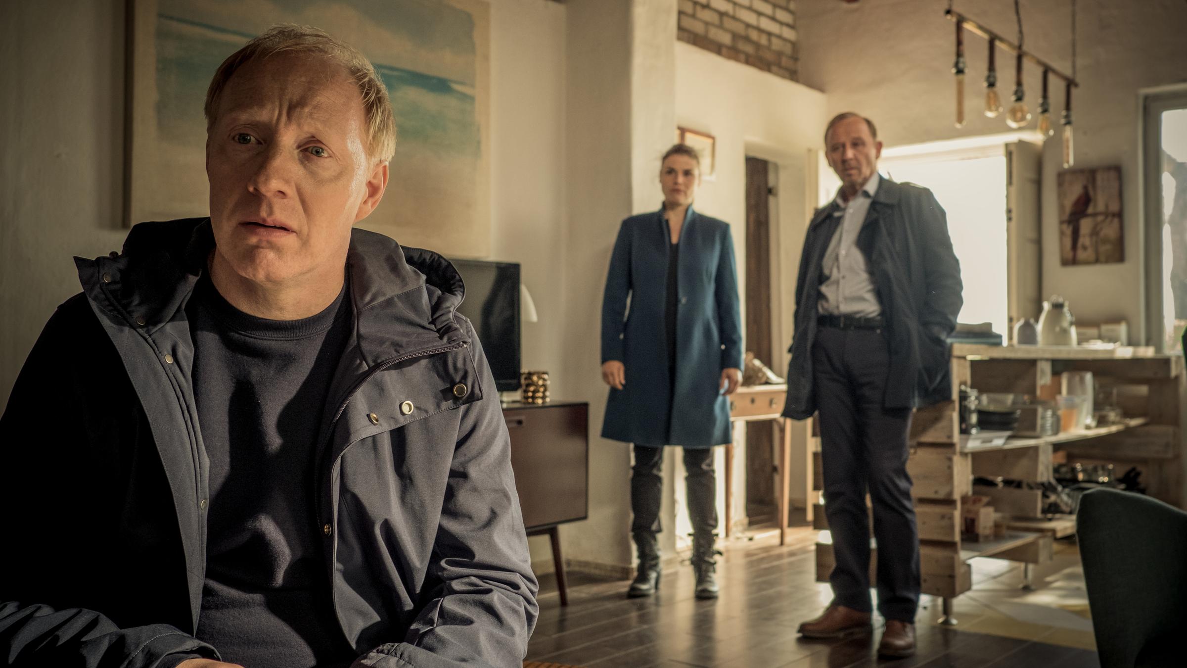 "Stralsund -  Doppelkopf": Peter Thies (Simon Schwarz) sitzt in einem Wohnzimmer und wird von Nina Petersen (Katharina Wackernagel) und Karl Hidde (Alexander Held), die hinter ihm stehen, beobachtet.