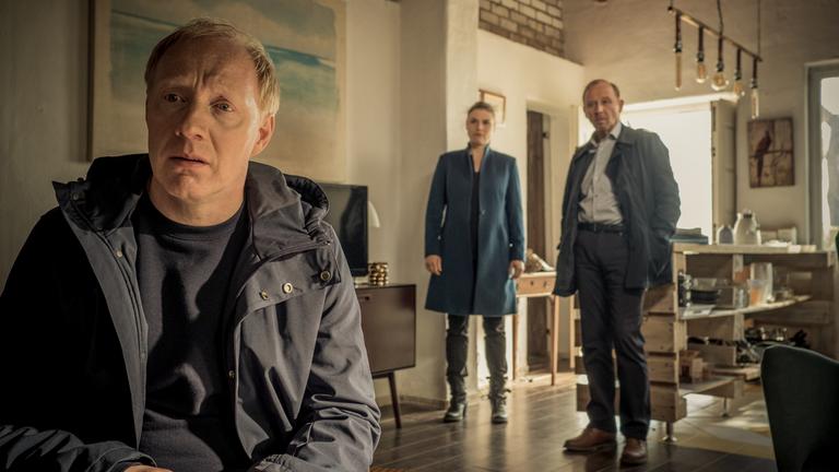 "Stralsund -  Doppelkopf": Peter Thies (Simon Schwarz) sitzt in einem Wohnzimmer und wird von Nina Petersen (Katharina Wackernagel) und Karl Hidde (Alexander Held), die hinter ihm stehen, beobachtet.
