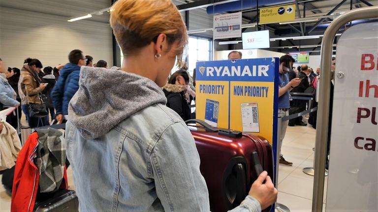 "ZDFzoom: Fliegen am Limit - der Preis des Wachstums": In einer Abflughalle steht eine Frau mit kurzen blonden Haaren und einem Koffer in der Hand an einer Gepäckabfertigungsstelle. Im Hintergrund mehrere Fluggäste beim Einchecken.