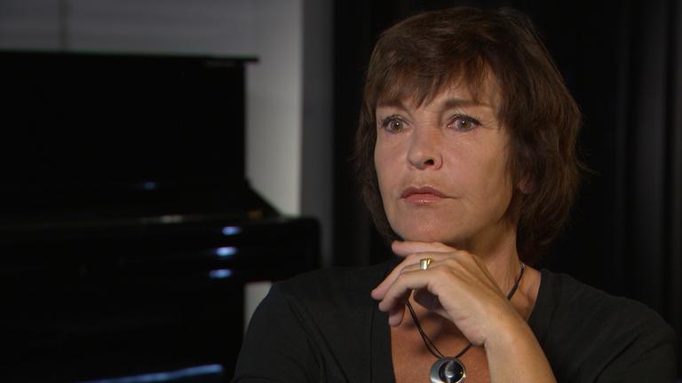 "Generationen-Wende": Katrin Sass sitzt vor schwarzem Hintergrund in Interviewsituation. Großaufnahme. Sie hält die Hand unter das Kinn, schaut fragend.