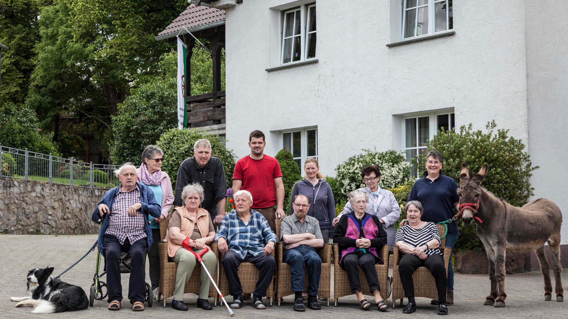 "37°: Bauernhof statt Altersheim - Eine Senioren-WG auf dem Land": Senioren sitzen auf Flechtstühlen während Familie Müller hinter den Senioren steht. Alle haben sich vor dem Wohnhaus versammelt.  Ganz rechts steht der Esel des Hofes. Links liegt ein Hund.