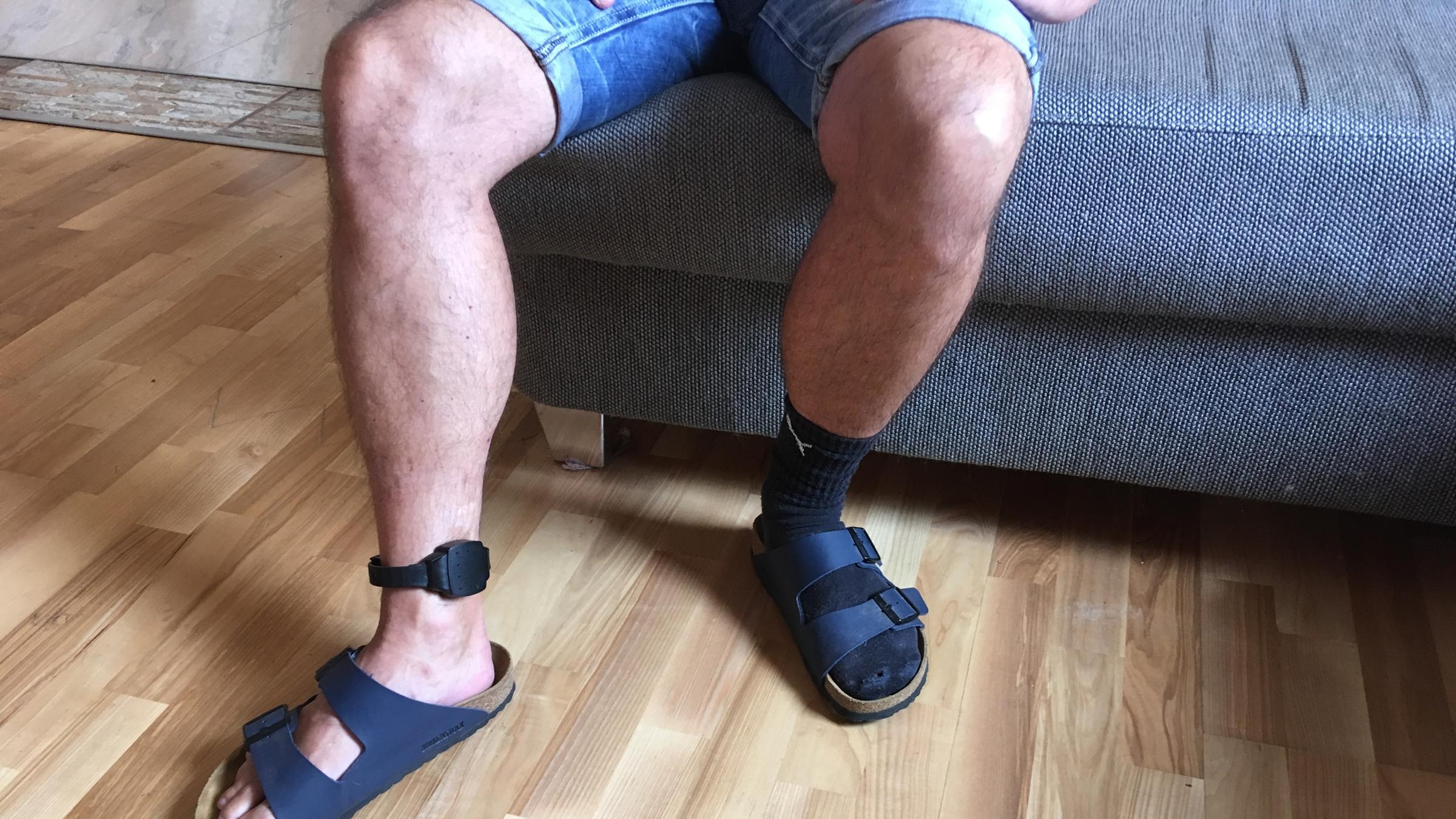"plan b - Knast ohne Gitter": Nacktes Bein eines Mannes, der auf einer Couch sitzt, mit elektronischer Fußfessel.