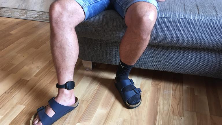 "plan b - Knast ohne Gitter": Nacktes Bein eines Mannes, der auf einer Couch sitzt, mit elektronischer Fußfessel.