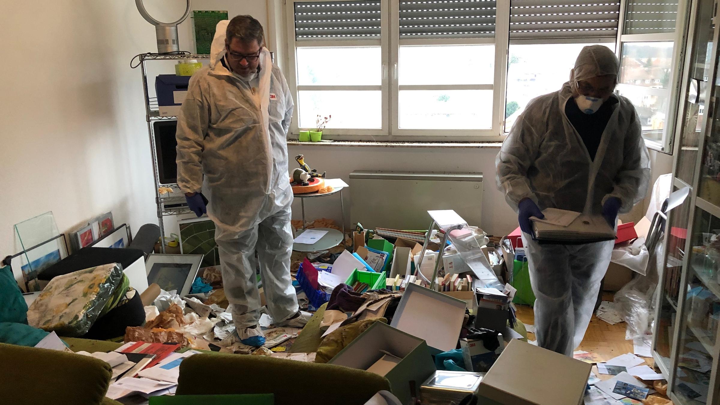 "Verwalter des einsamen Todes": Ein heller Raum mit großem Fenster, bei dem die Jalousie zu einem Drittel heruntergelassen wurde. Auf dem Boden verstreut Unmengen von Bildern, Papieren, Kartons und weiterer Unrat. Dazwischen zwei Männer in transparenten weißen Schutzanzügen.