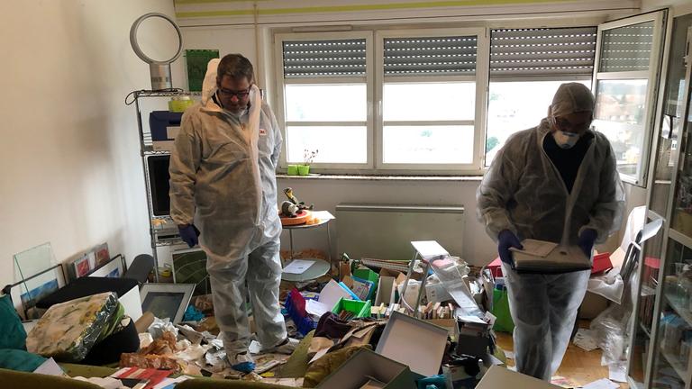 "Verwalter des einsamen Todes": Ein heller Raum mit großem Fenster, bei dem die Jalousie zu einem Drittel heruntergelassen wurde. Auf dem Boden verstreut Unmengen von Bildern, Papieren, Kartons und weiterer Unrat. Dazwischen zwei Männer in transparenten weißen Schutzanzügen.