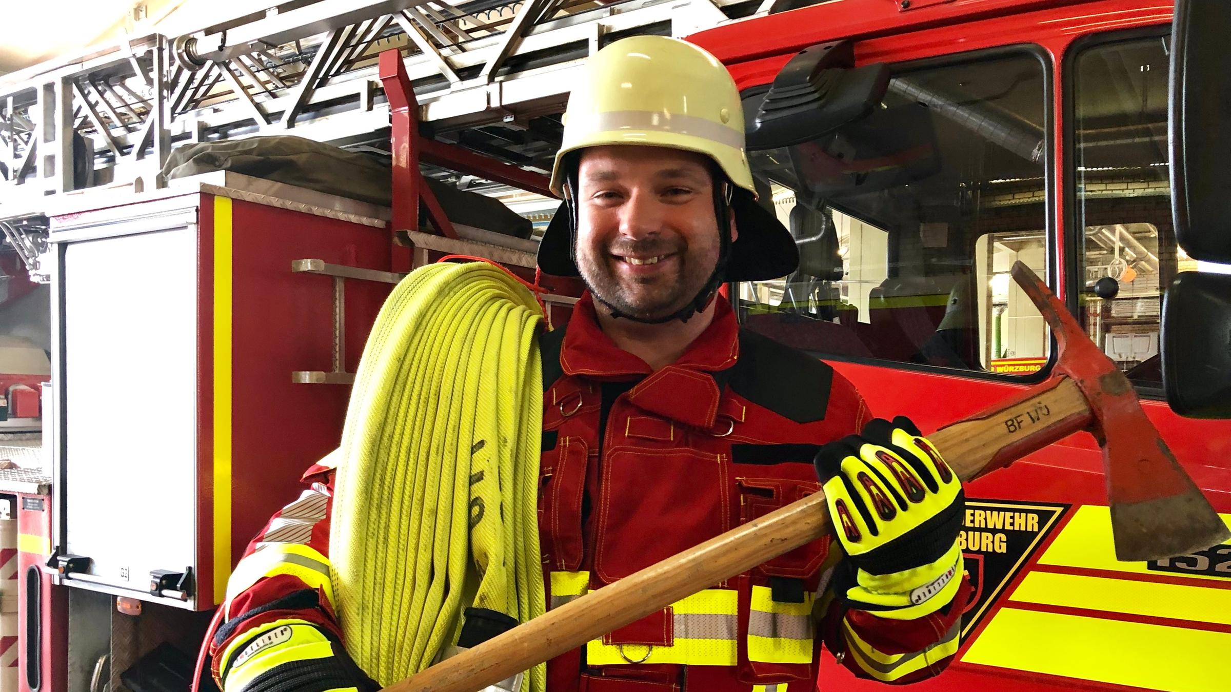 "Ein guter Grund zu feiern - Allerheiligen mit Pater Nikodemus Schnabel": Pater Nikodemus Schnabel trägt eine rote Feuerwehruniform einen gelben Helm. In den Händen hält er eine große Axt und über der linken Schulter hängt einer gelber Feuerwehrschlauch. Hinter ihm ist eine Feuerwehrlöschfahrzeug zu sehen.