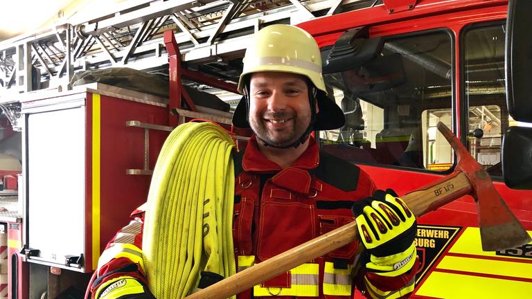 "Ein guter Grund zu feiern - Allerheiligen mit Pater Nikodemus Schnabel": Pater Nikodemus Schnabel trägt eine rote Feuerwehruniform einen gelben Helm. In den Händen hält er eine große Axt und über der linken Schulter hängt einer gelber Feuerwehrschlauch. Hinter ihm ist eine Feuerwehrlöschfahrzeug zu sehen.