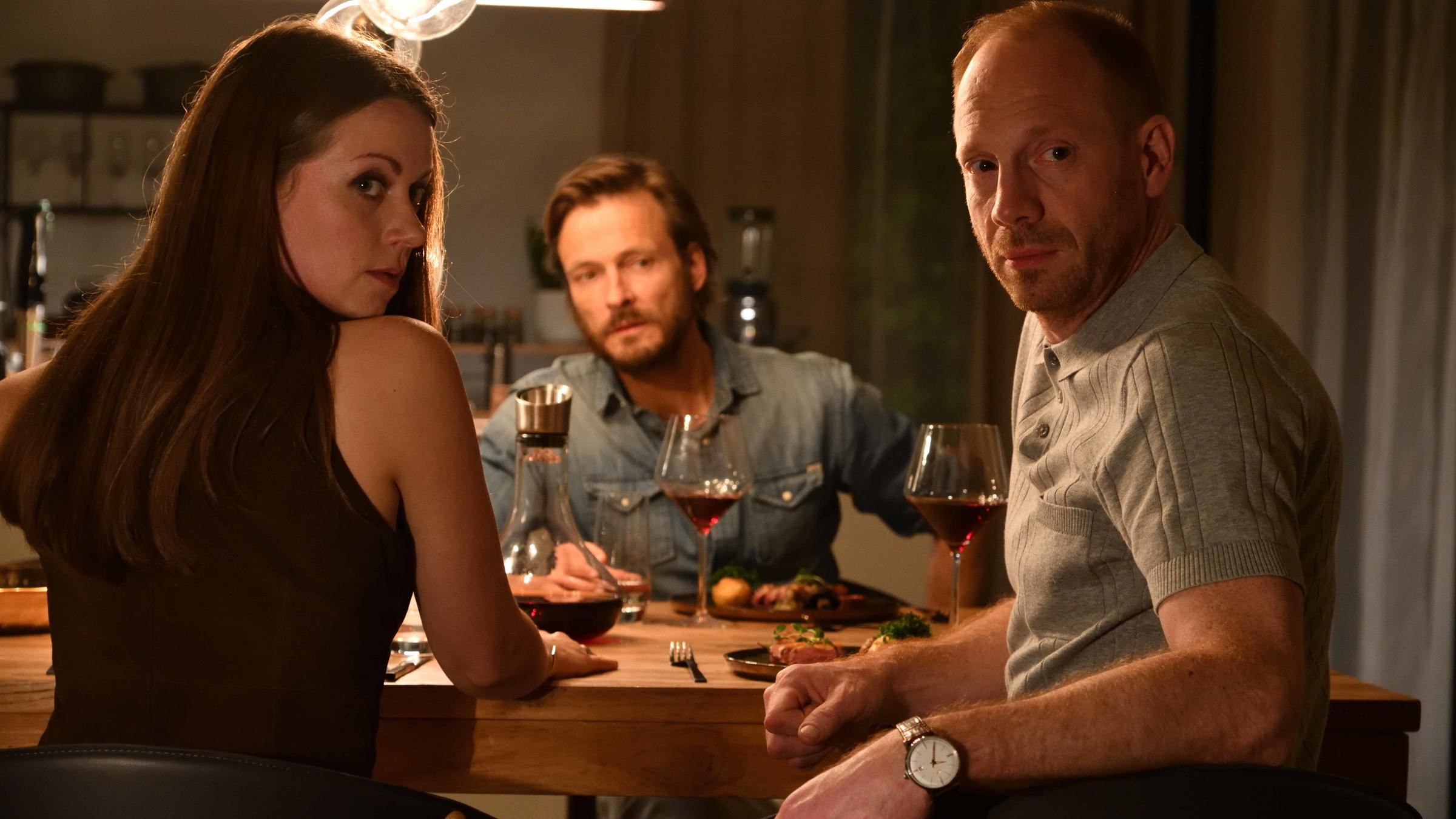 „Herr und Frau Bulle – Totentanz“: Yvonne (Alice Dwyer) und Heiko Wills (Johann von Bülow) essen gemeinsam mit Heikos Freund Piet Bernsdorff (Andreas Pietschmann) zu Abend. Auf dem Tisch stehen Rotweingläser, Teller und ein Dekanter. Alle drei schauen erschrocken in dieselbe Richtung.