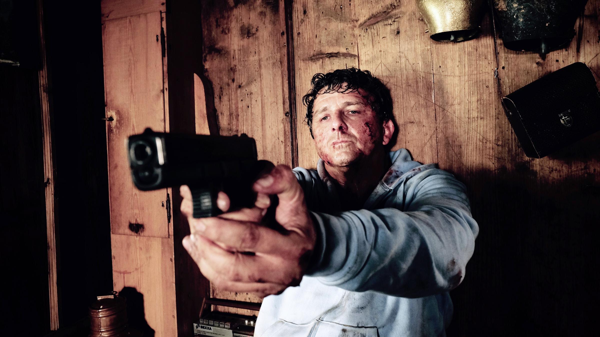 "Flucht durchs Höllental": Rechtsanwalt Klaus Burg (Hans Sigl) sitzt verletzt mit gezogener Waffe vor einer Holzbretterwand, an der Kuhglocken befestigt sind und eine Umhängetasche hängt. Neben ihm auf der Bank steht ein Kassettenrekorder und in der Ecke eine Milchkanne.