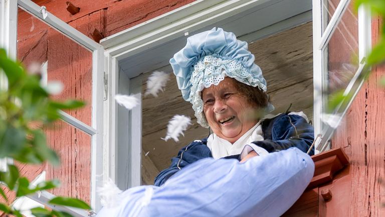 "Terra X: Magie der Märchen - Frau Holle und ihre versunkene Welt": Frau Holle schüttelt ein Kissen aus dem Fenster heraus. Federn fliegen umher.