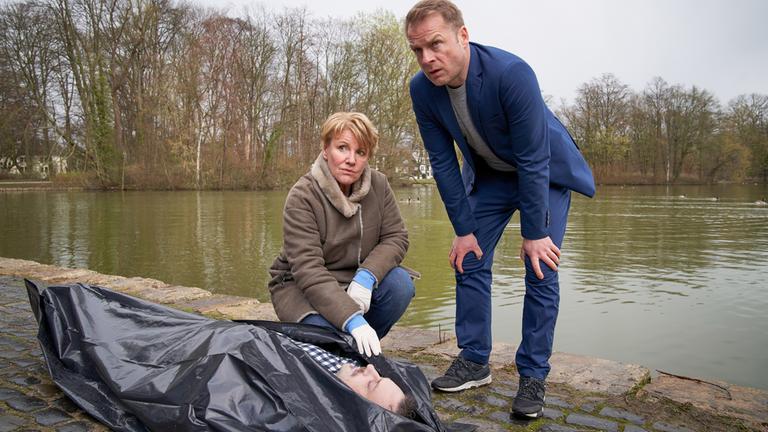 "Marie Brandt und die Liebe zu viert": Am Ufer liegt eine Leiche im Leichensack. Marie Brand (Mariele Millowitsch) sitzt in der Hocke daneben. Neben ihr steht leicht gebeugt Jürgen Simmel (Hinnerk Schönemann). Beide schauen beide über die Leiche hinweg in die gleiche Richtung.