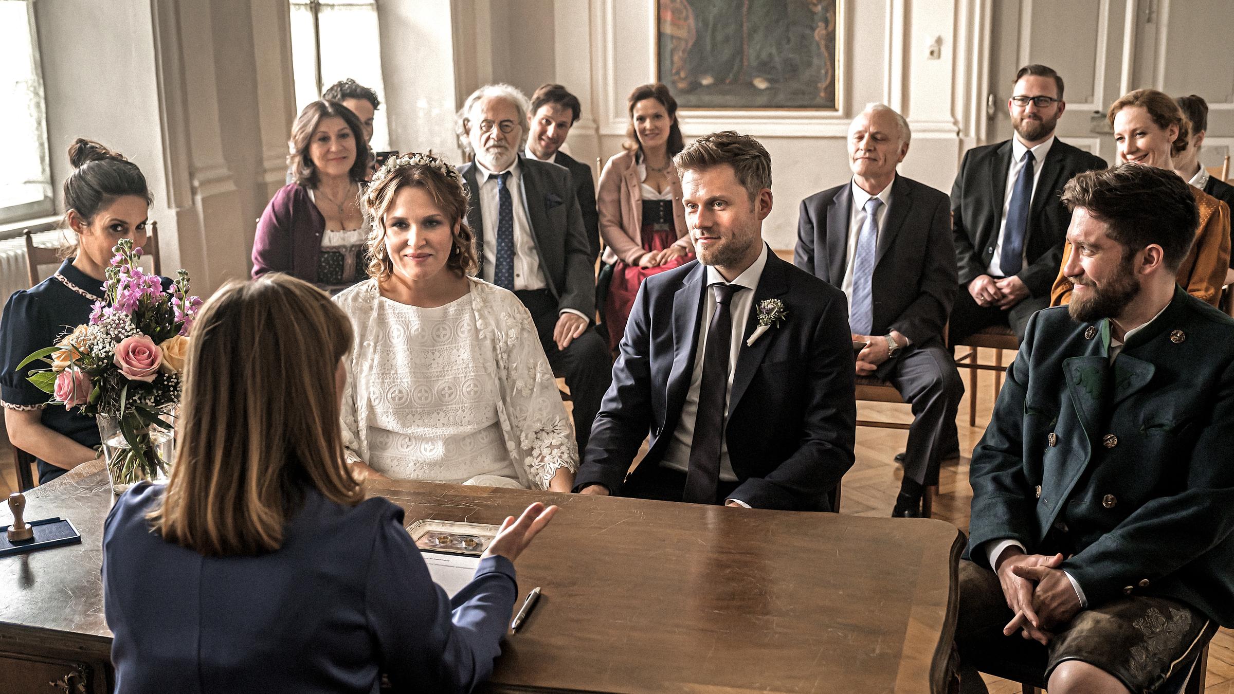 "Lena Lorenz - Außergewöhnlich einzigartig": Das Brautpaar Lena Lorenz (Judith Hoersch) und Quirin Pankofer (Jens Atzorn) sitzen an einem antiken Holztisch. Ihnen gegenüber sitzt die Standesbeamtin (Gabrielle Odinis), welche gestikulierend zu ihnen spricht. Um die beiden herum sitzen Familie und Freunde. Alle hören lächelnd zu.