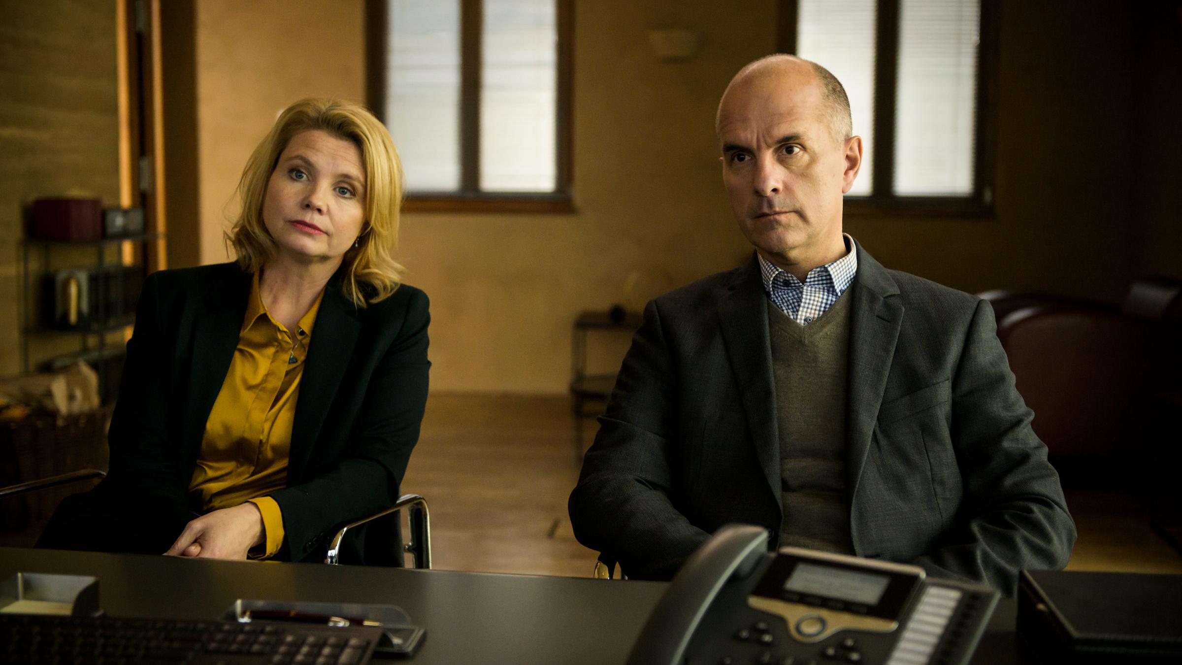 "Merz gegen Merz – Der Vertrag": Vor einem schwarzen Bürotisch sitzen Anne Merz (Annette Frier) und Erik Merz (Christoph Maria Herbst) nebeneinander und sehen ihr Gegenüber aufmerksam an.