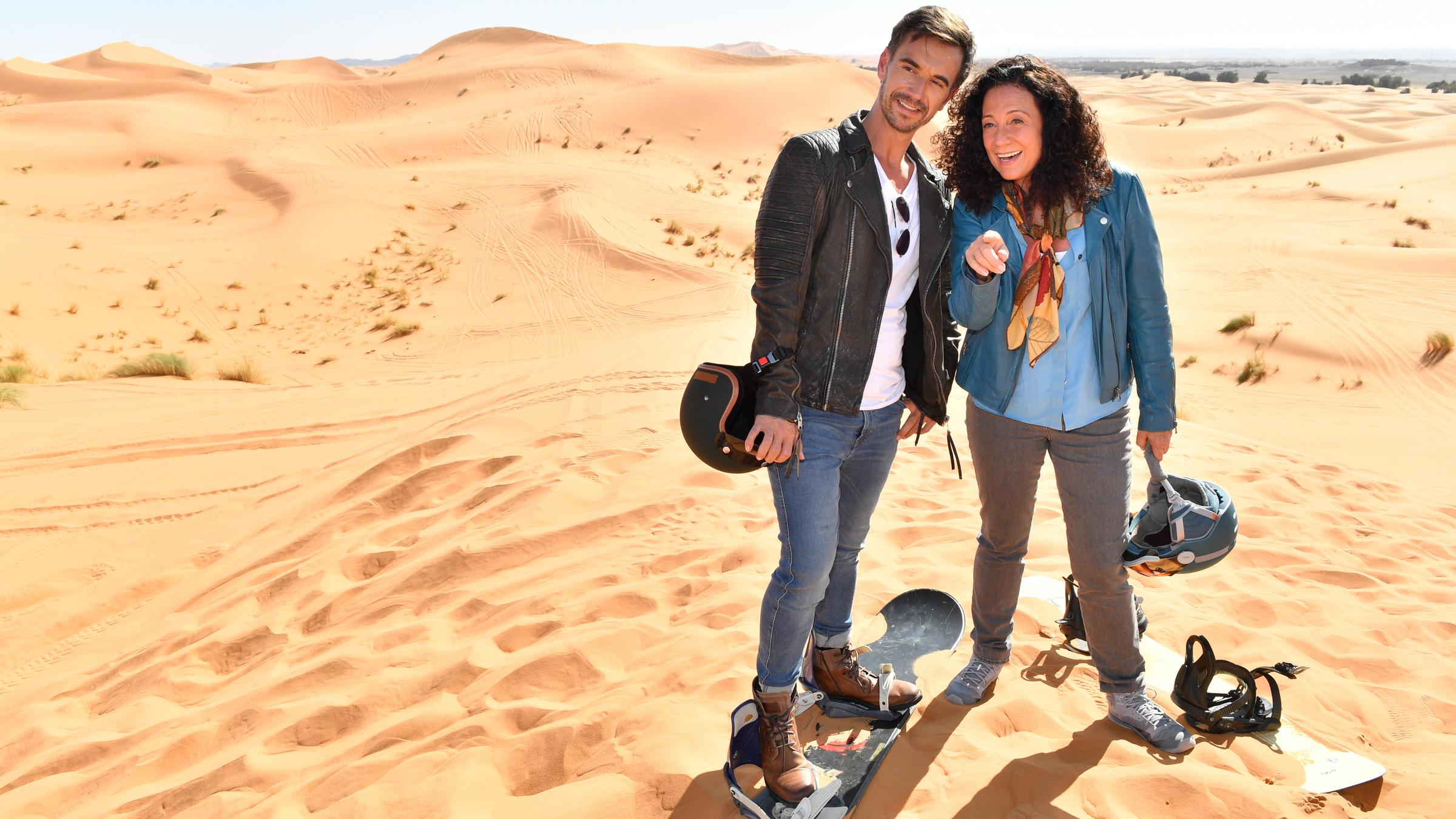"Das Traumschiff: Marokko": Max Parger (Florian Silbereisen) und Hanna Liebhold (Barbara Wussow) stehen mit Snowboards und Helmen auf einer Düne in der Wüste. Hanna zeigt auf etwas und die beiden sehen freudig in die gleiche Richtung.