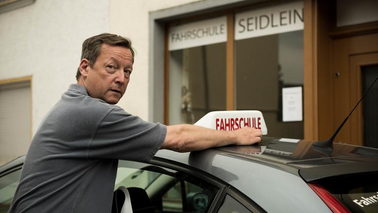 "Wir wären andere Menschen": Rupert (Matthias Brandt) setzt das Fahrschulschild auf das Dach vom Auto.