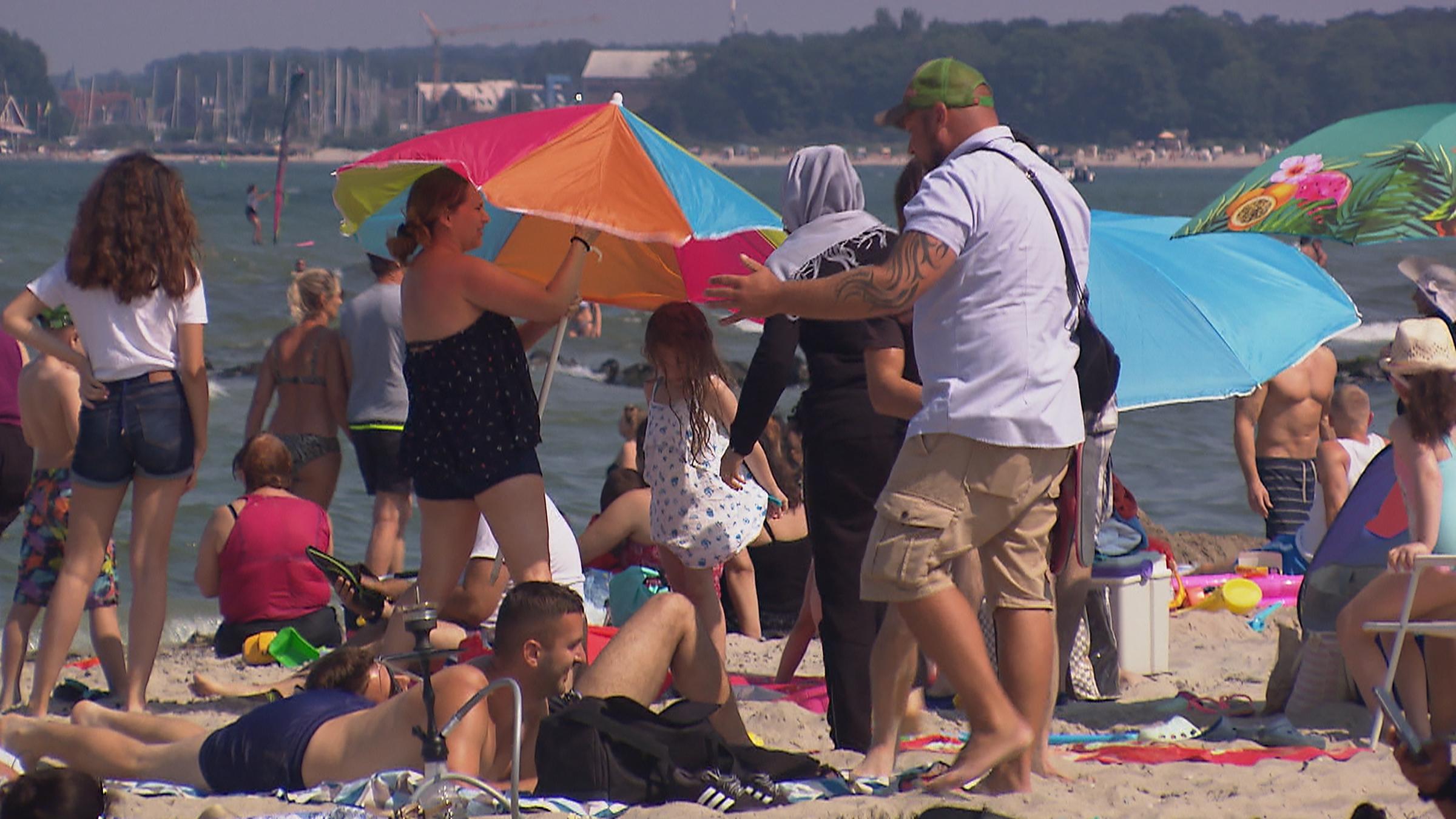 "ZDF.reportage: Sommer, Sonne, Sicherheit - Strandleben in Corona-Zeiten": Viele Badegäste tummeln sich am Timmendorfer Strand. Mehrere Sonnenschirme sind aufgespannt. Ein Mitarbeiter der Kurverwaltung in einem weißem T-Shirt spricht mit den Sonnenanbetern.