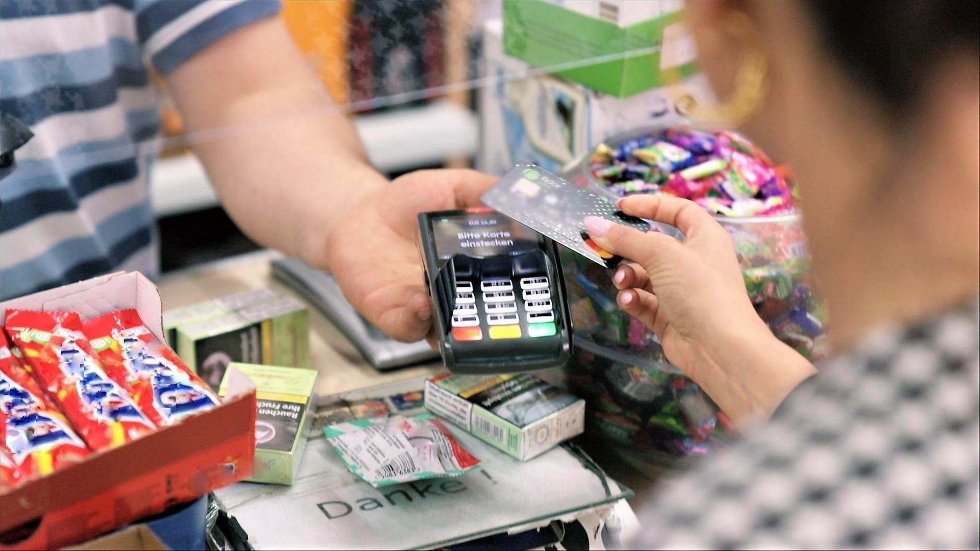 ZDFzoom: Karte oder Cash": Eine Hand hält ein Kartenlesegerät über einen Verkaufstisch eines Kiosks. Im Vordergrund sieht man von hinten eine Frau, die sich mit ihrer Kreditkarte dem Kartenlesegerät nähert.