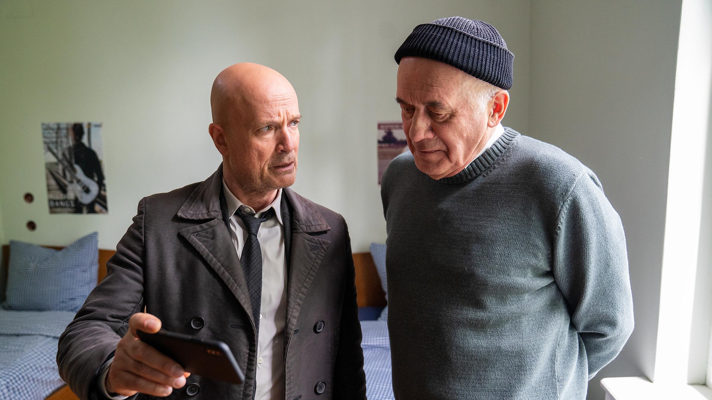 "Der Kriminalist – Das Böse in uns": Bruno Schumann (Christian Berkel) und der Sozialarbeiter Günther Ellis (Hanns Zischler) stehen in einem Heimzimmer nebeneinander. Auf seinem Handy zeigt Schumann Ellis ein Video und beobachtet seine Reaktion.